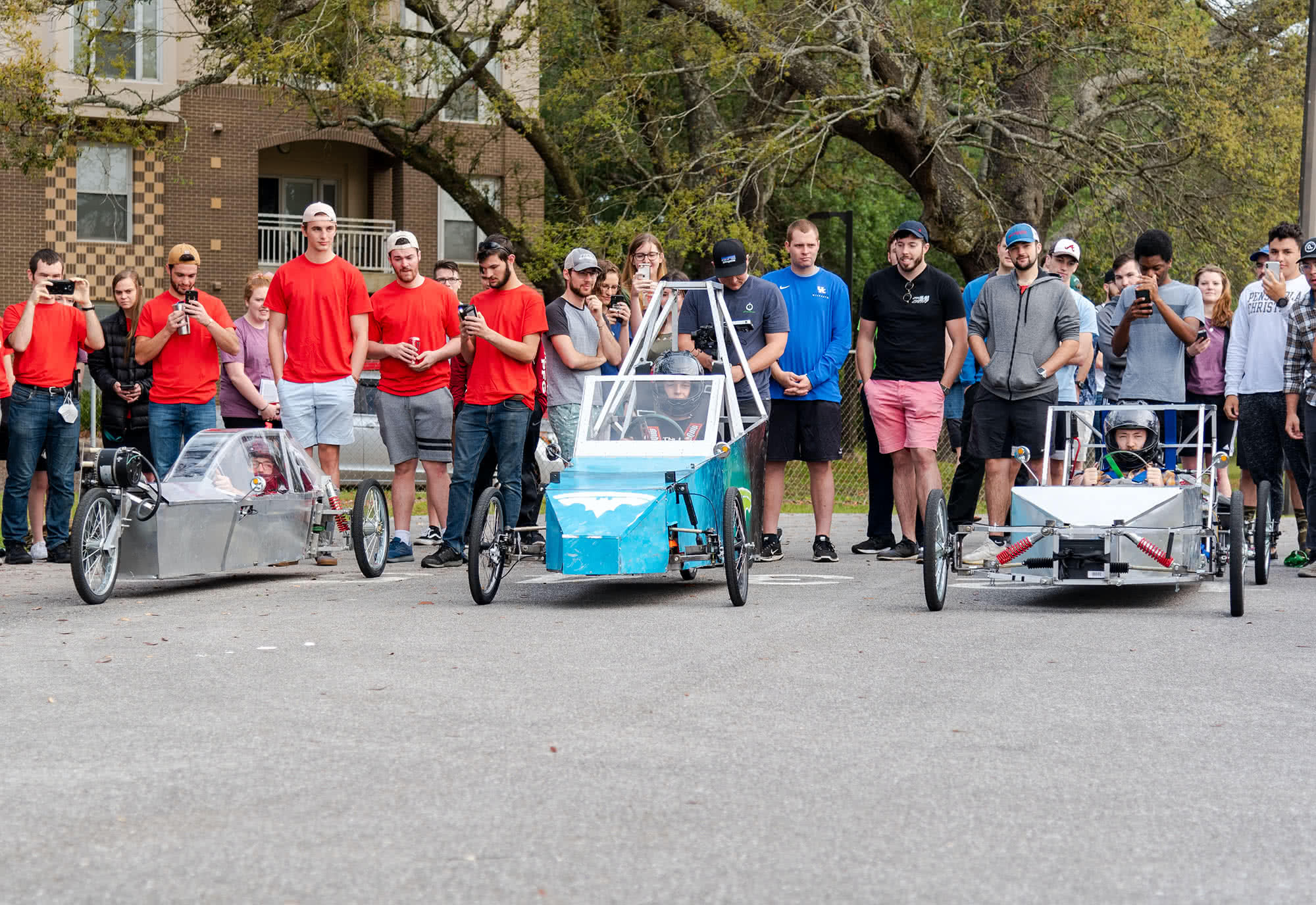 Students gather around 3 students in electric vehicles preparing to begin a race. 