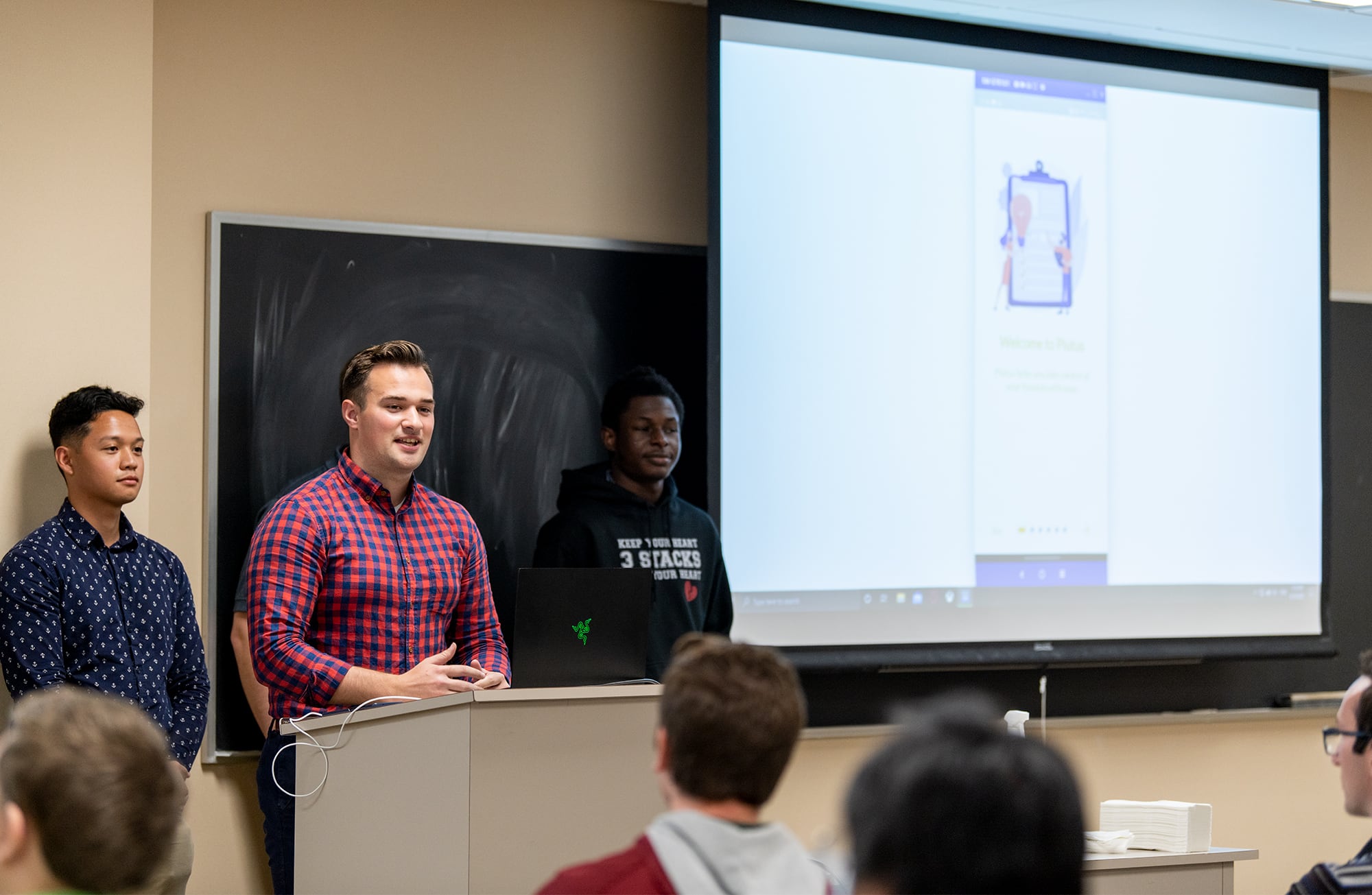 Three male students standing in front of a class of students and presenting.