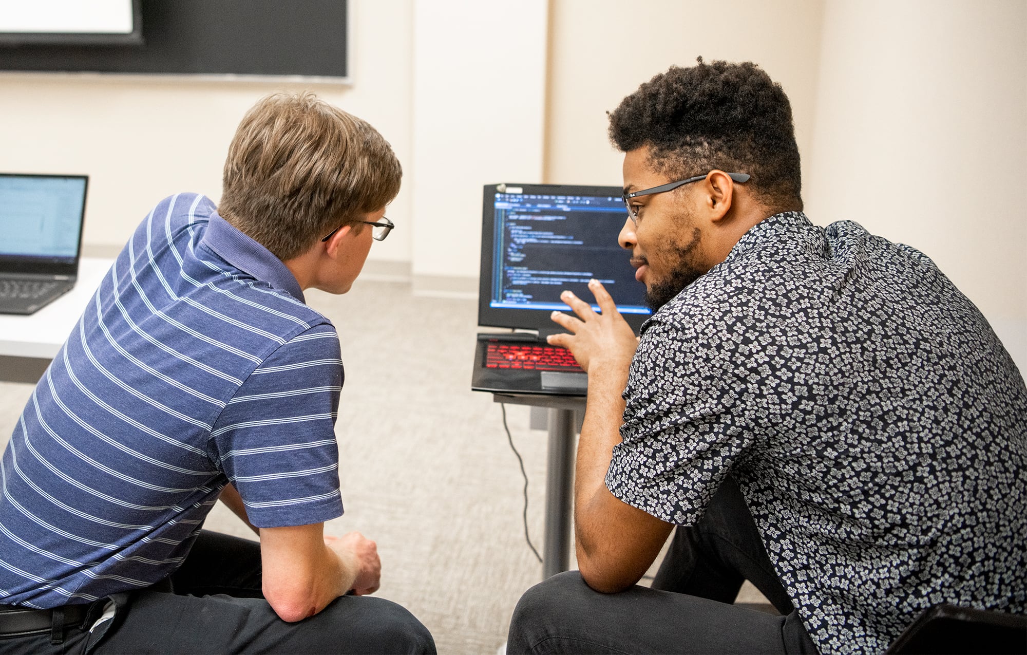 A male student explaining software to another male student at a laptop.