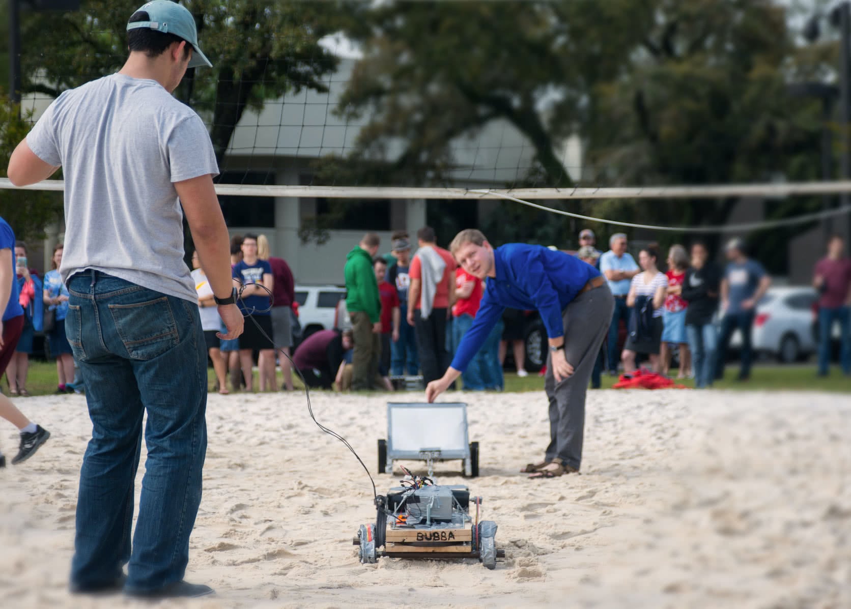Two male engineering students standing with their robots facing each other on the sand volleyball court.