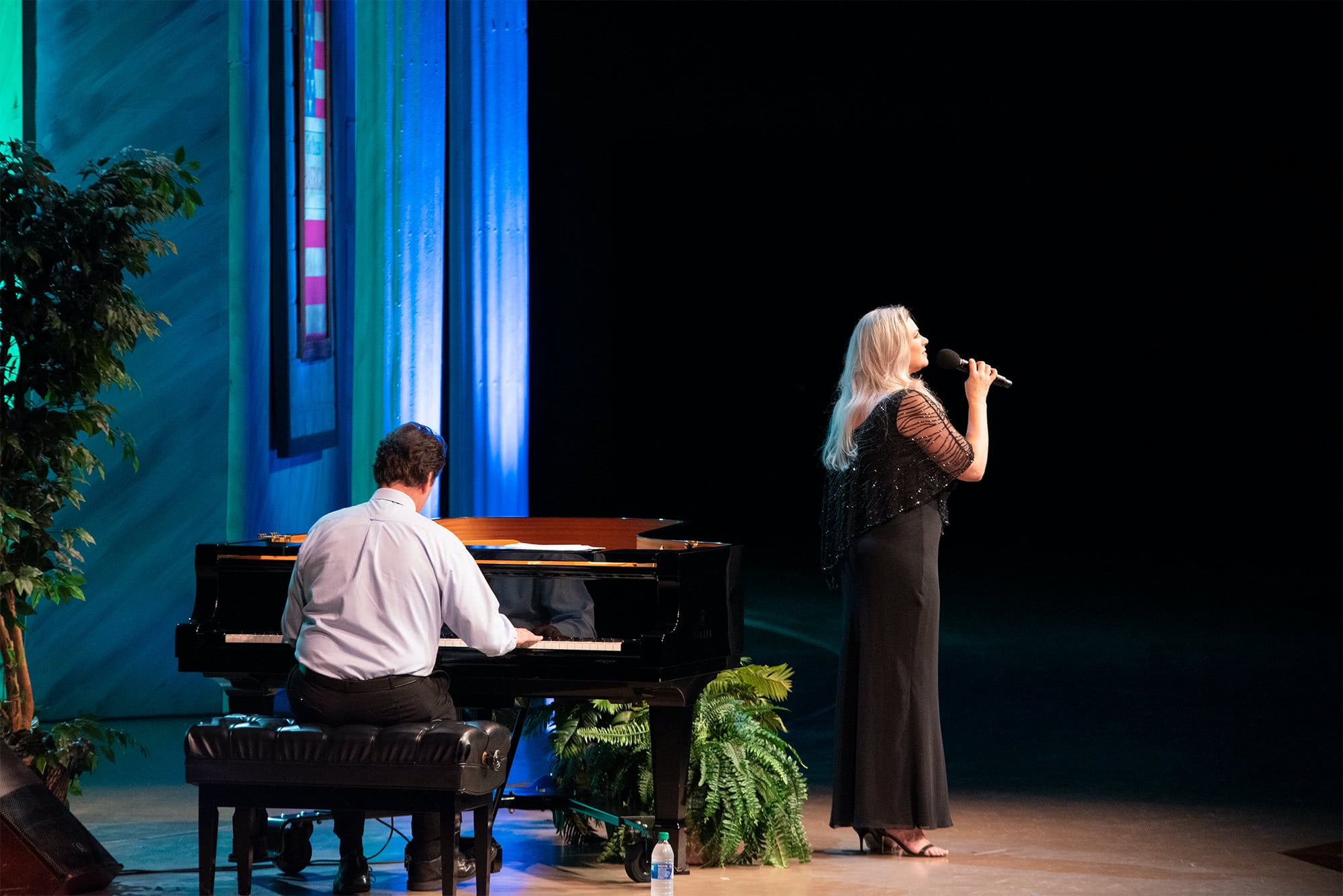 Female Liberty Voices singer wearing a black dress and singing next to a man playing the piano.