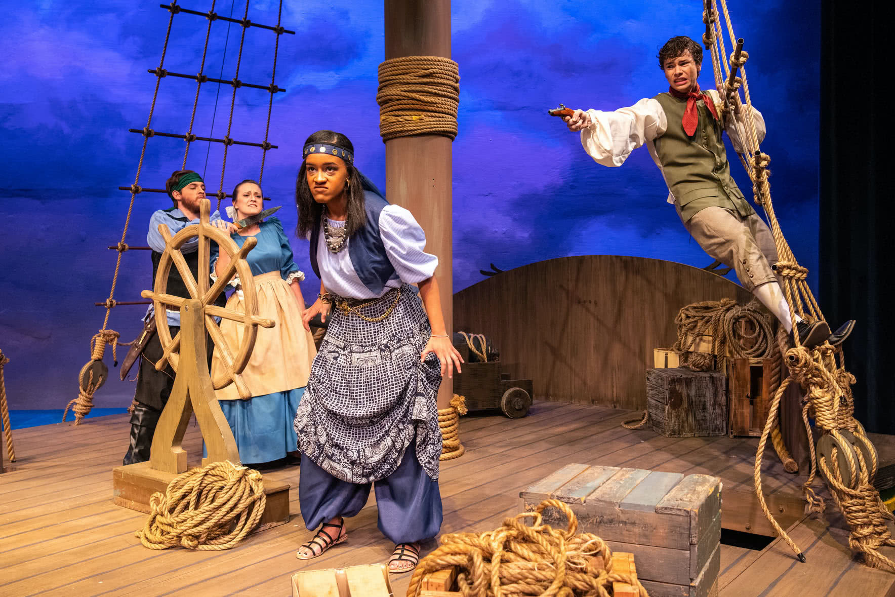 A female pirate looks angry on board a pirate ship, behind her a boy aims a pistol at her; in the background another pirate threatens a lady with a knife in PCC's Treasure Island.