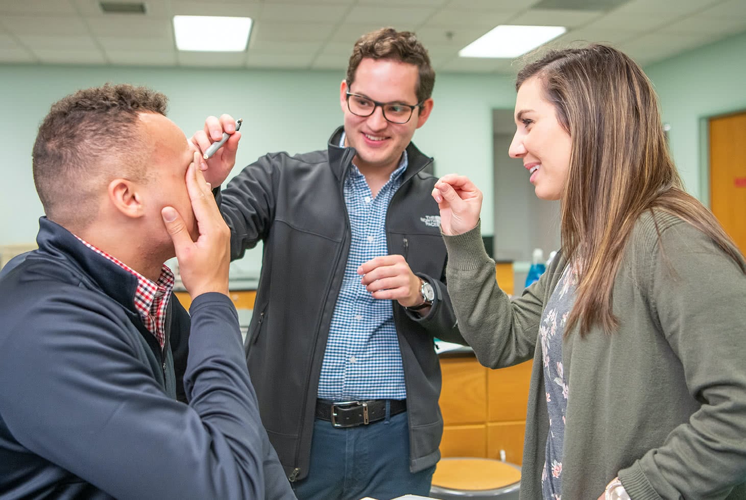 A male student covering his eye while two other students hold a light and examine his other eye at the start of spring semester classes.