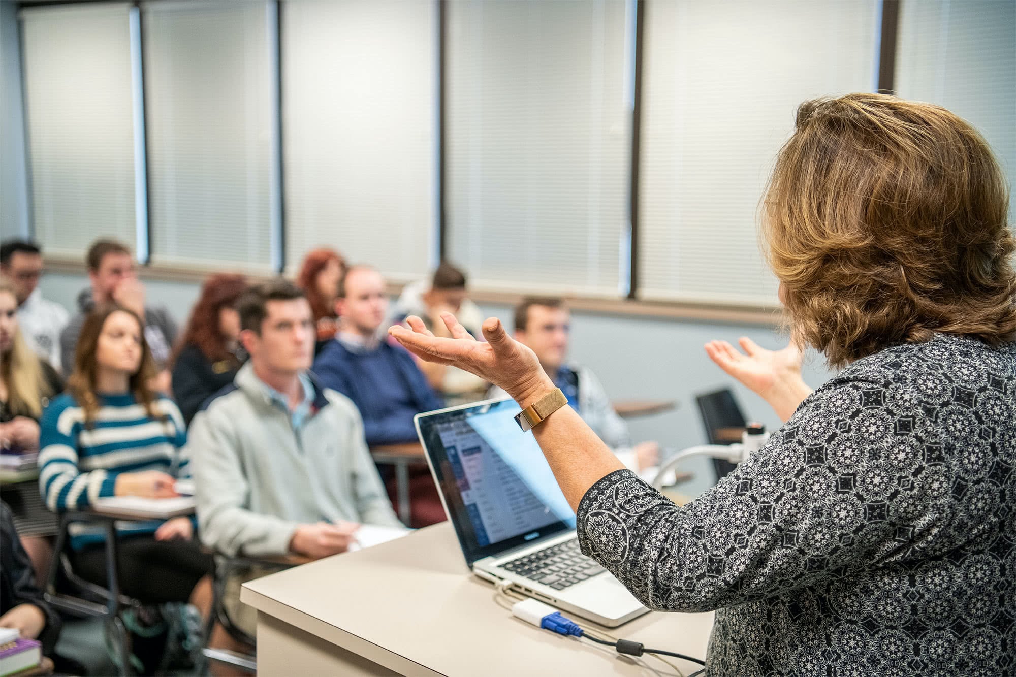 Female teacher standing at classroom podium and holding her hands upwards.