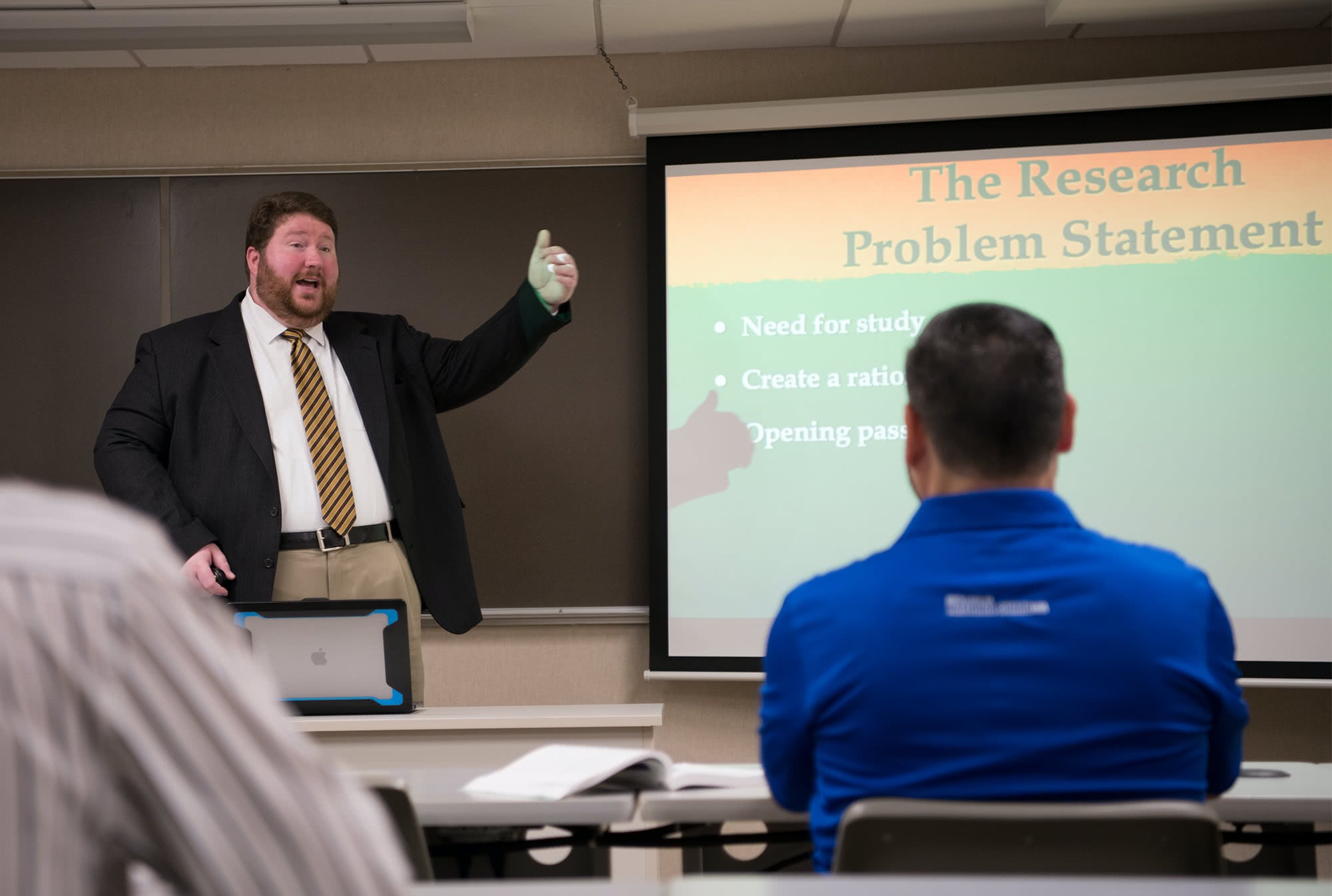 Professor standing at the front of a classroom and lecturing to graduate students. 