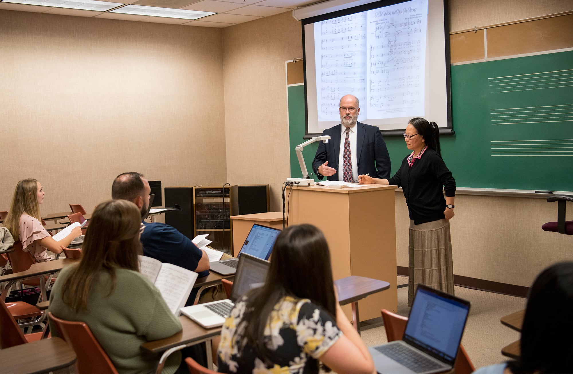 Two professors standing at the front of a classroom and instructing a class of graduate students. 