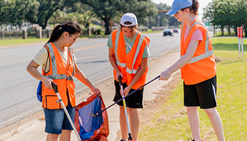 Three students in neon orange vests pick up trash along a highway.
